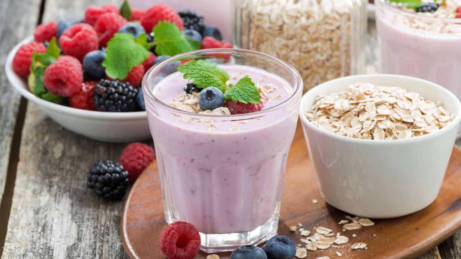 A glass of berry smoothie topped with oats and berries, garnished with mint, is on a wooden tray. Next to it is a bowl of oats and a bowl of assorted berries, including raspberries, blueberries, and blackberries.