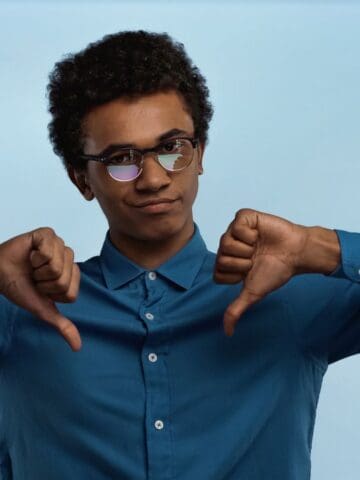 A person wearing glasses and a blue shirt stands against a light blue background. They have a neutral expression and are giving a double thumbs-down gesture with both hands.