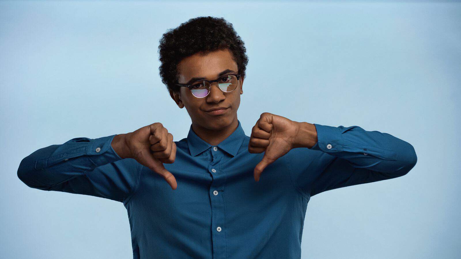 A person wearing glasses and a blue shirt stands against a light blue background. They have a neutral expression and are giving a double thumbs-down gesture with both hands.