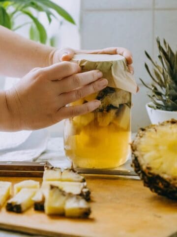 A person places a paper cover on a jar filled with pineapple pieces and liquid. A half pineapple and sliced pieces are on a cutting board with a knife. A pineapple top and potted plant are in the background on a kitchen counter.