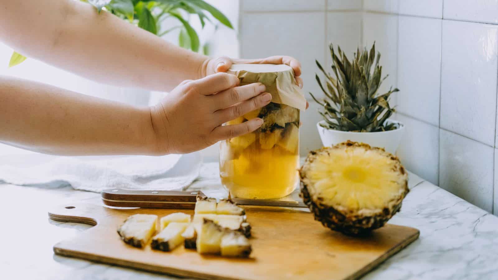 A person places a paper cover on a jar filled with pineapple pieces and liquid. A half pineapple and sliced pieces are on a cutting board with a knife. A pineapple top and potted plant are in the background on a kitchen counter.