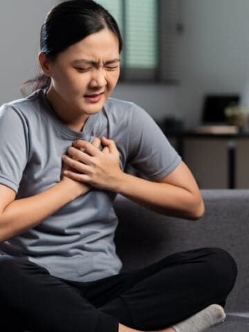A woman sitting cross-legged on a gray couch holds her chest with both hands, appearing to be in pain. She wears a gray T-shirt and black pants. A guitar, plant, and desk are in the blurred background.