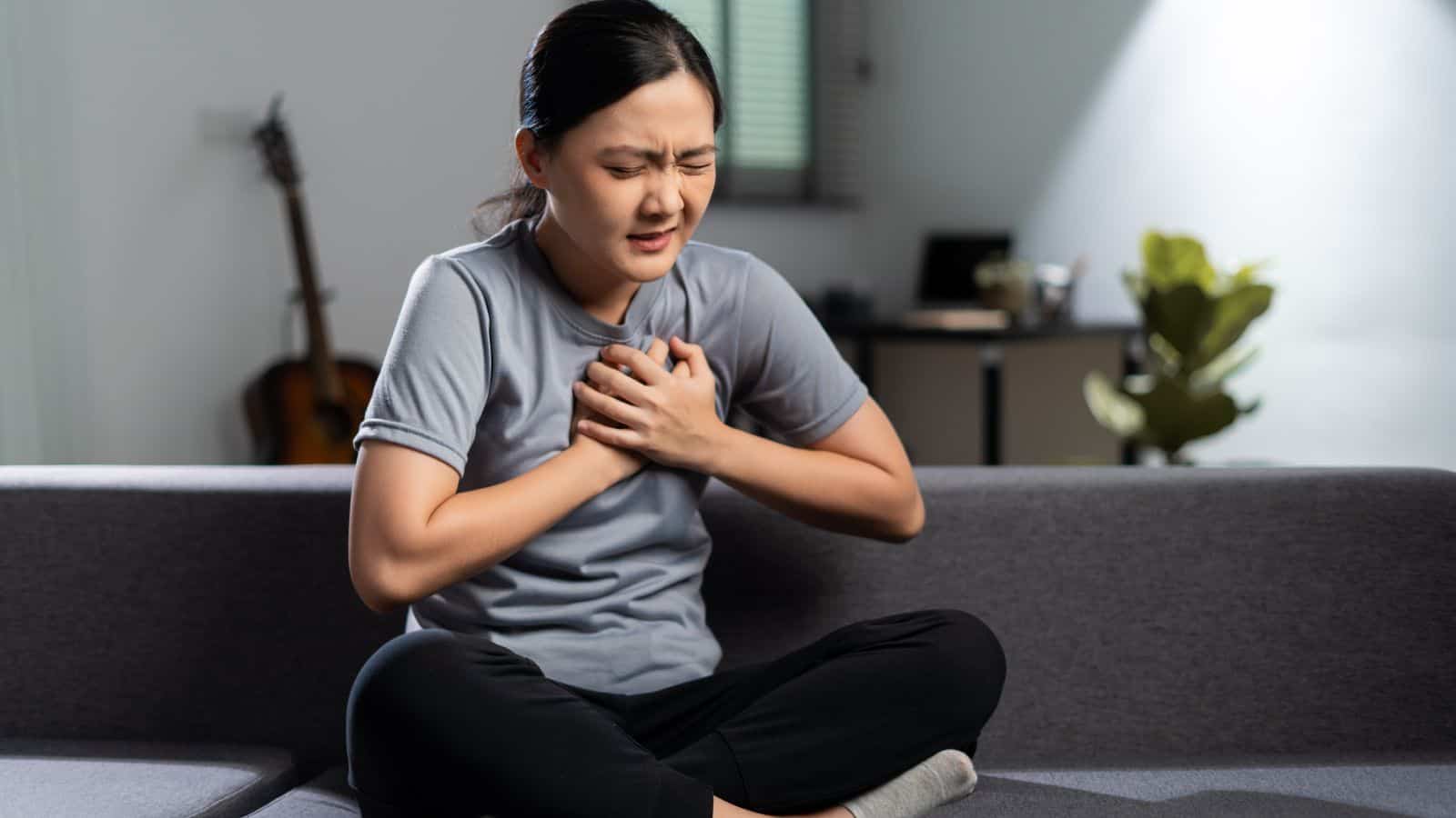 A woman sitting cross-legged on a gray couch holds her chest with both hands, appearing to be in pain. She wears a gray T-shirt and black pants. A guitar, plant, and desk are in the blurred background.
