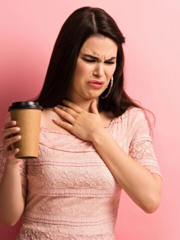 A woman in a pink dress holds a paper coffee cup in her left hand while touching her throat with her right hand. Her expression appears uncomfortable. The background is a solid pink color.