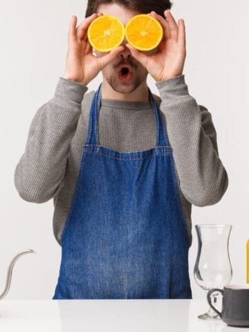 A person in a gray shirt and blue apron holds orange halves over their eyes while standing at a counter. The counter has various items, including a coffee pot, a glass, a jar of coffee beans, a cup, and a bottle of orange juice.