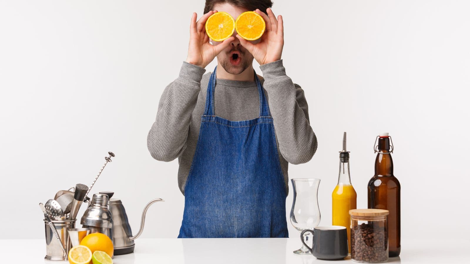 A person in a gray shirt and blue apron holds orange halves over their eyes while standing at a counter. The counter has various items, including a coffee pot, a glass, a jar of coffee beans, a cup, and a bottle of orange juice.