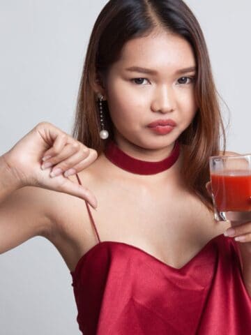 A woman in a red dress holds a glass of red juice in one hand while giving a thumbs-down gesture with the other. She has long brown hair and is wearing pearl earrings. Her expression is neutral. The background is plain gray.