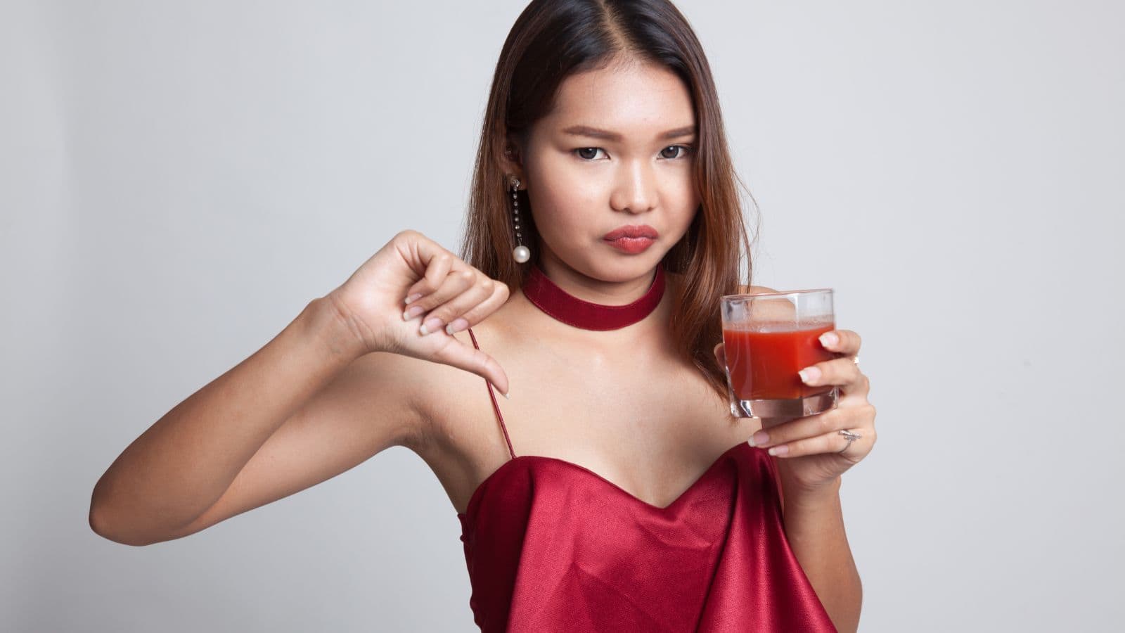 A woman in a red dress holds a glass of red juice in one hand while giving a thumbs-down gesture with the other. She has long brown hair and is wearing pearl earrings. Her expression is neutral. The background is plain gray.