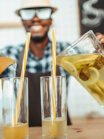 A person wearing a hat and eyeglasses is pouring drinks from a pitcher into two glasses. One pitcher contains yellow liquid and the other contains a light green liquid with visible lemon slices. A menu board is blurred in the background.