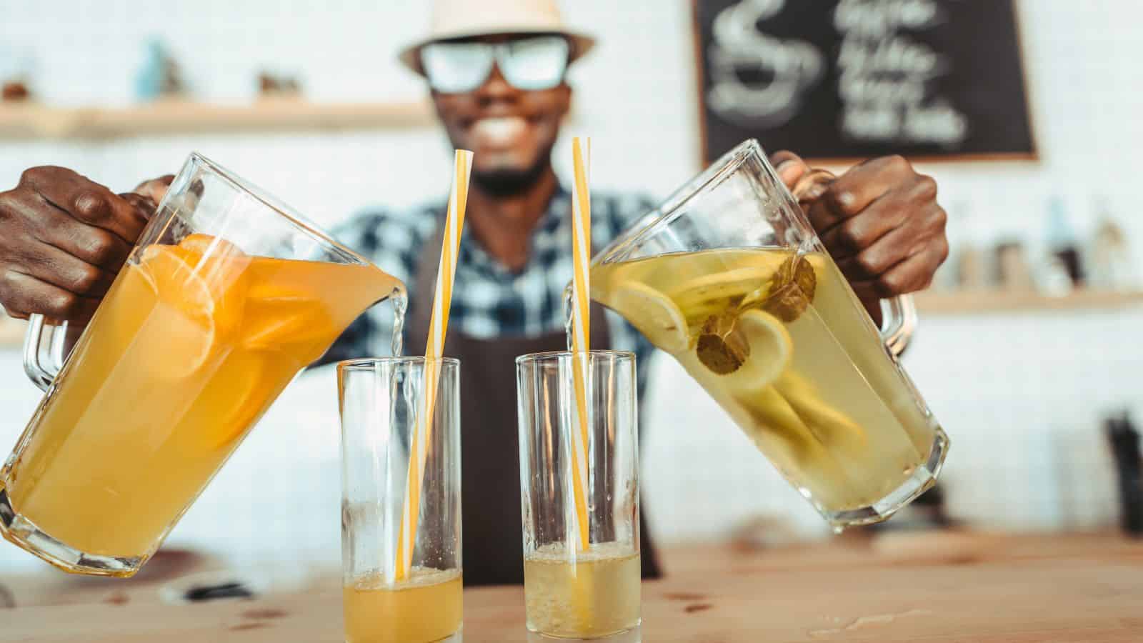 A person wearing glasses and a checkered shirt is pouring drinks into two tall glasses from pitchers. One pitcher contains orange juice, and the other has a lemon and lime beverage. The person is smiling, with a cheerful demeanor.