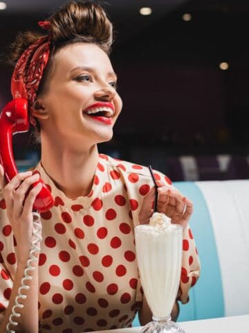 A woman with a retro hairstyle and red polka dot blouse is holding a red rotary phone and smiling. She is sitting at a diner booth with a milkshake in front of her. The background is dimly lit.
