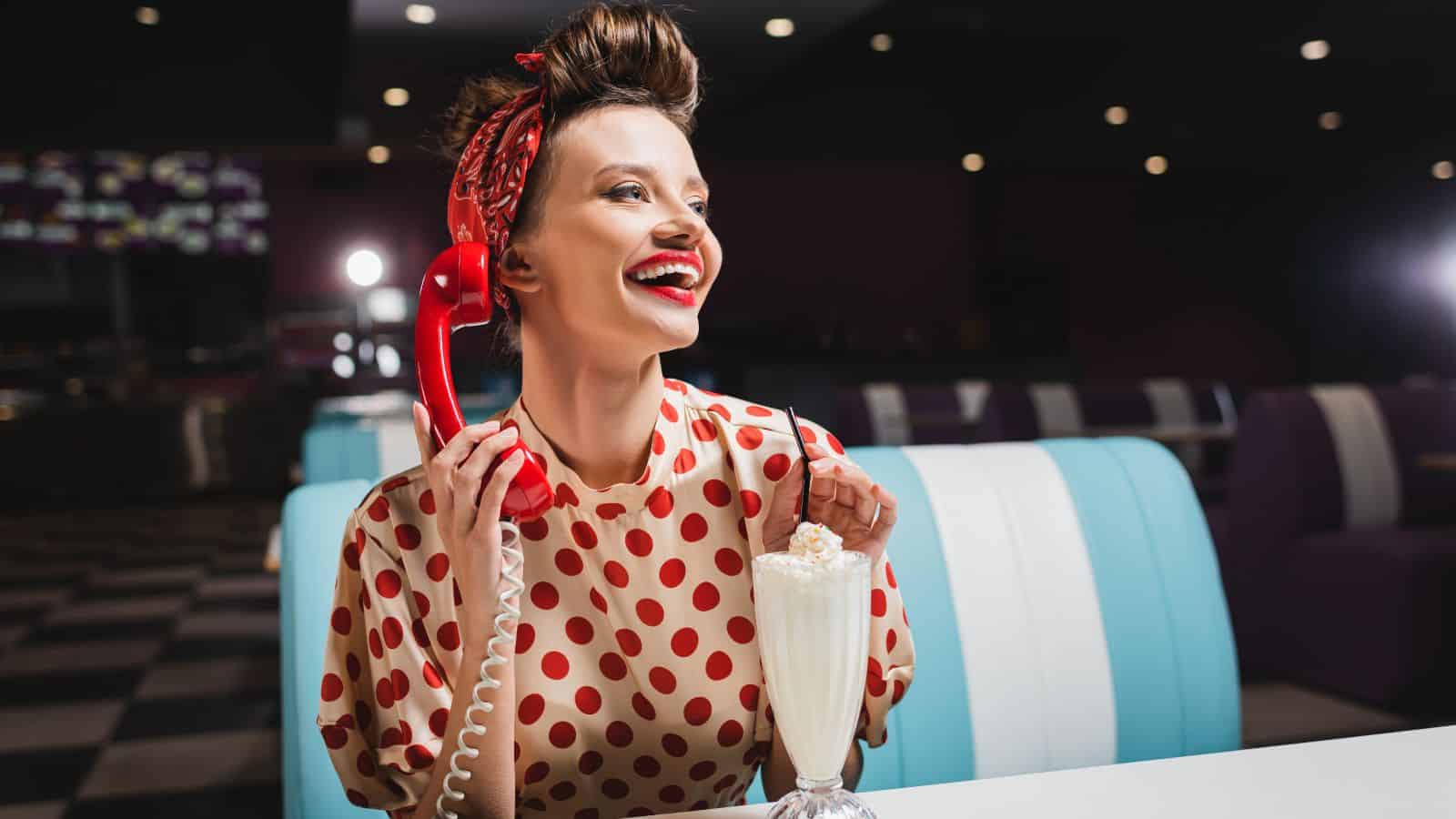 A woman with a retro hairstyle and red polka dot blouse is holding a red rotary phone and smiling. She is sitting at a diner booth with a milkshake in front of her. The background is dimly lit.