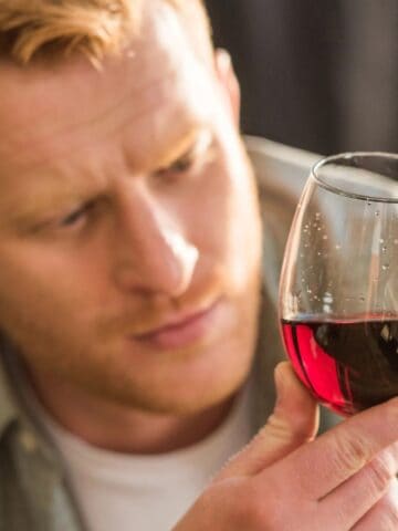 A person with light brown hair, wearing a green checkered shirt, is holding a glass of red wine. The individual is looking intently at the liquid against a blurred background.