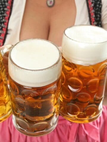 A person wearing traditional attire holds three large glass mugs filled with beer. Each mug has a thick layer of foam on top. The background features a blue and white checkered pattern.