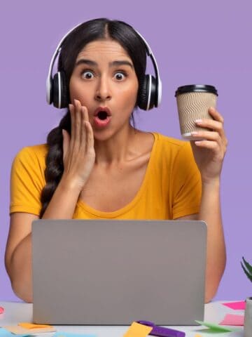 A woman in a yellow shirt sits at a desk with a laptop. She wears headphones and holds a coffee cup, looking surprised. The desk has books, notepads, an apple, sticky notes, a plant, and a pen holder. The background is purple.