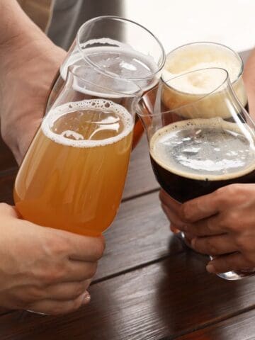 Four people clink glasses filled with different types of beer over a wooden table. The hands are visible, and each glass contains a distinct color and level of froth, indicating a variety of beers.