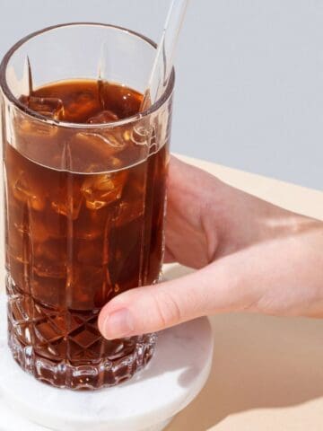 A hand holds a glass filled with iced coffee and a clear straw. The glass is positioned on a round, white coaster on a beige surface. A single coffee bean is resting on the coaster beside the glass.