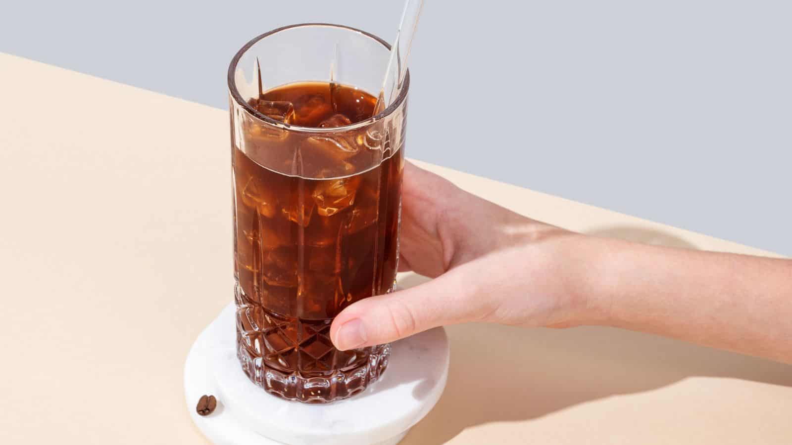 A hand holds a glass filled with iced coffee and a clear straw. The glass is positioned on a round, white coaster on a beige surface. A single coffee bean is resting on the coaster beside the glass.