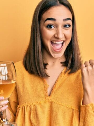 A person with shoulder-length brown hair is smiling widely and pointing with their thumb to the side. They are holding a large glass of a light beverage and are wearing a mustard yellow shirt. The background is a solid yellow color.