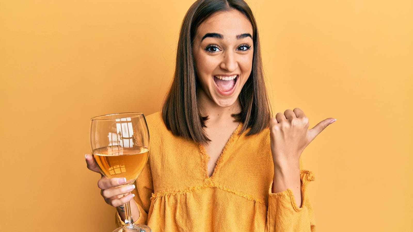 A person with shoulder-length brown hair is smiling widely and pointing with their thumb to the side. They are holding a large glass of a light beverage and are wearing a mustard yellow shirt. The background is a solid yellow color.