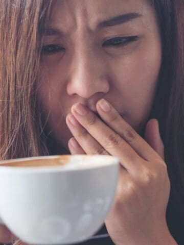 A person with long hair and wearing a striped shirt covers their mouth with a hand while holding a white cup of coffee. They have a concerned expression, set against a blurred background.