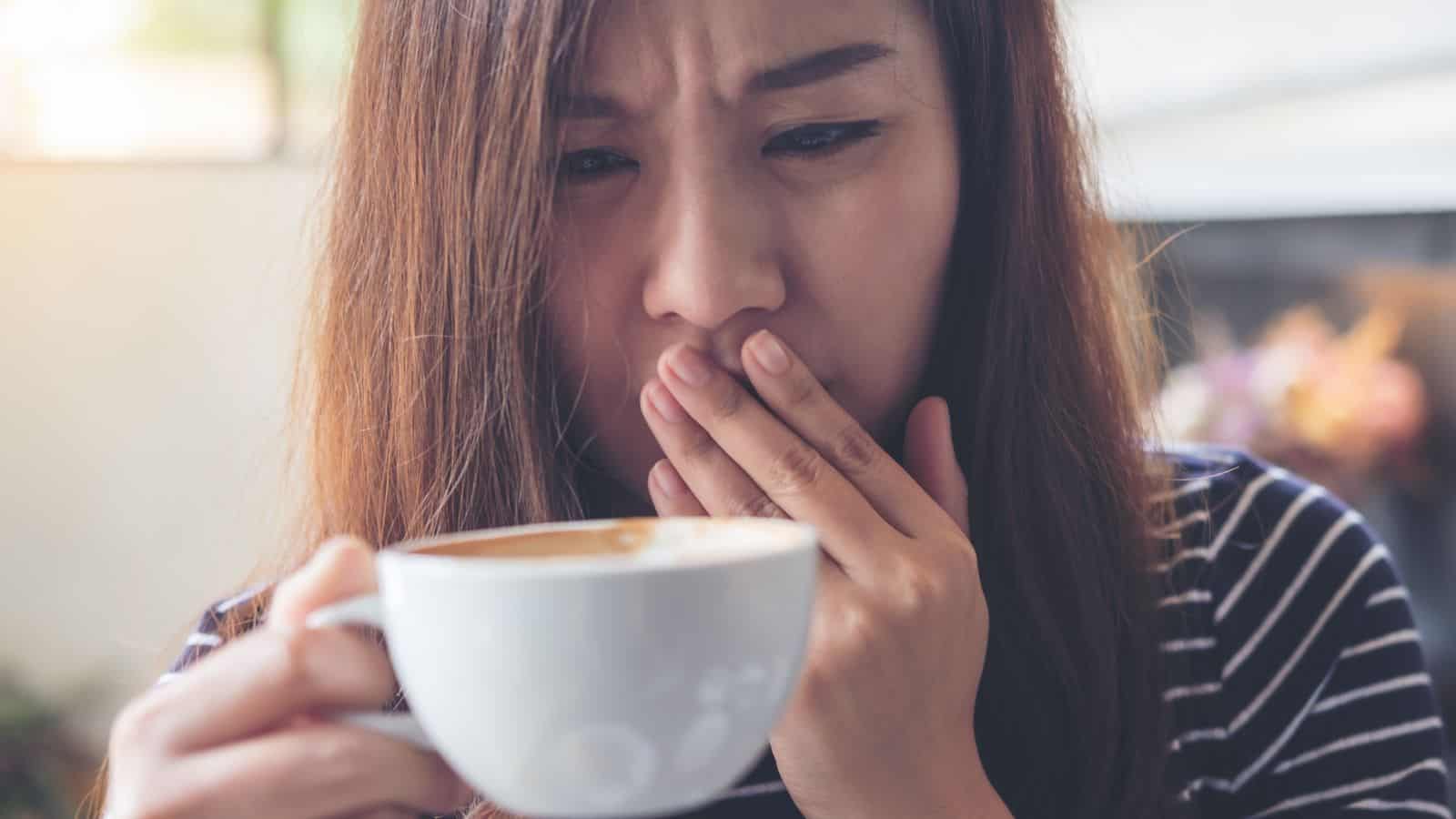 A person with long hair and wearing a striped shirt covers their mouth with a hand while holding a white cup of coffee. They have a concerned expression, set against a blurred background.