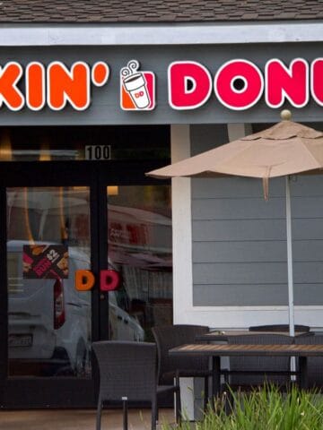 Exterior of a Dunkin' Donuts store featuring a sign with the brand’s logo above the entrance. Outside, there is a patio table with four chairs and an umbrella. The building has gray siding and a stone accent wall.