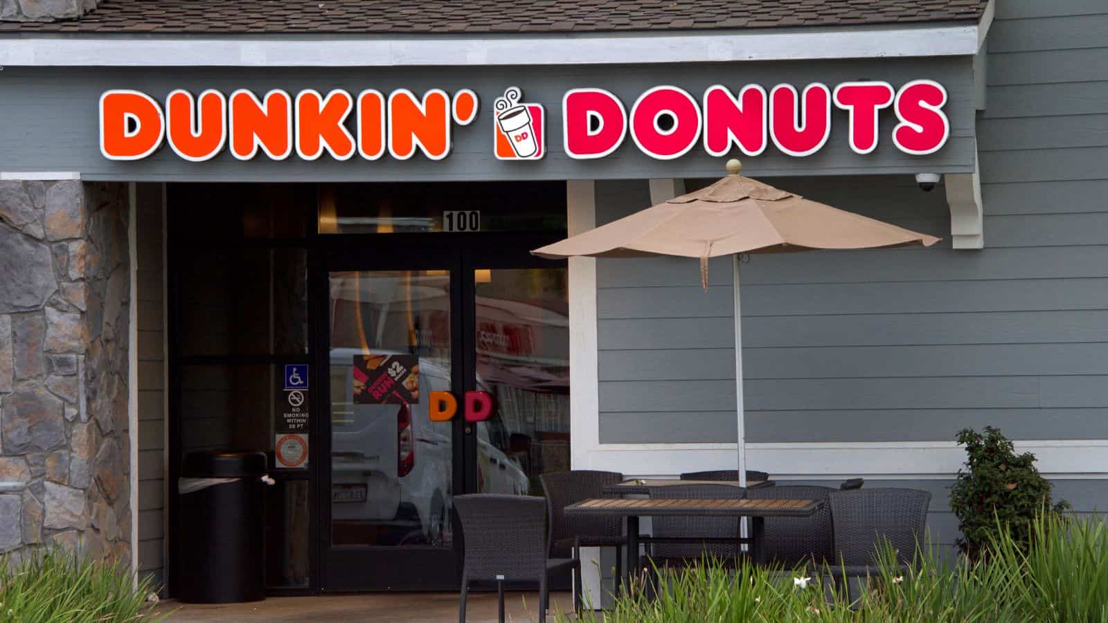 Exterior of a Dunkin' Donuts store featuring a sign with the brand&rsquo;s logo above the entrance. Outside, there is a patio table with four chairs and an umbrella. The building has gray siding and a stone accent wall.
