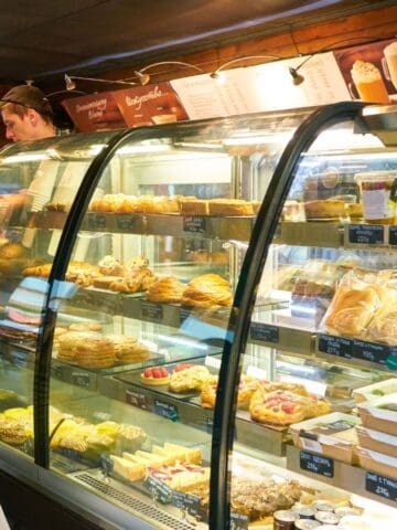 A man stands in front of a glass display case filled with various pastries and baked goods inside a bakery. An employee is visible behind the counter. The display has labels and a menu board overhead. The man holds a white plastic bag.