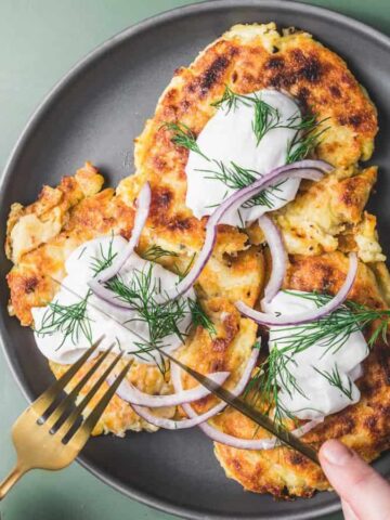 A plate with three golden-brown potato pancakes topped with sour cream, dill, and red onion slices. A hand holds a fork and knife, poised to cut into one pancake. A small bowl of red onion slices is on the side. The background is green.