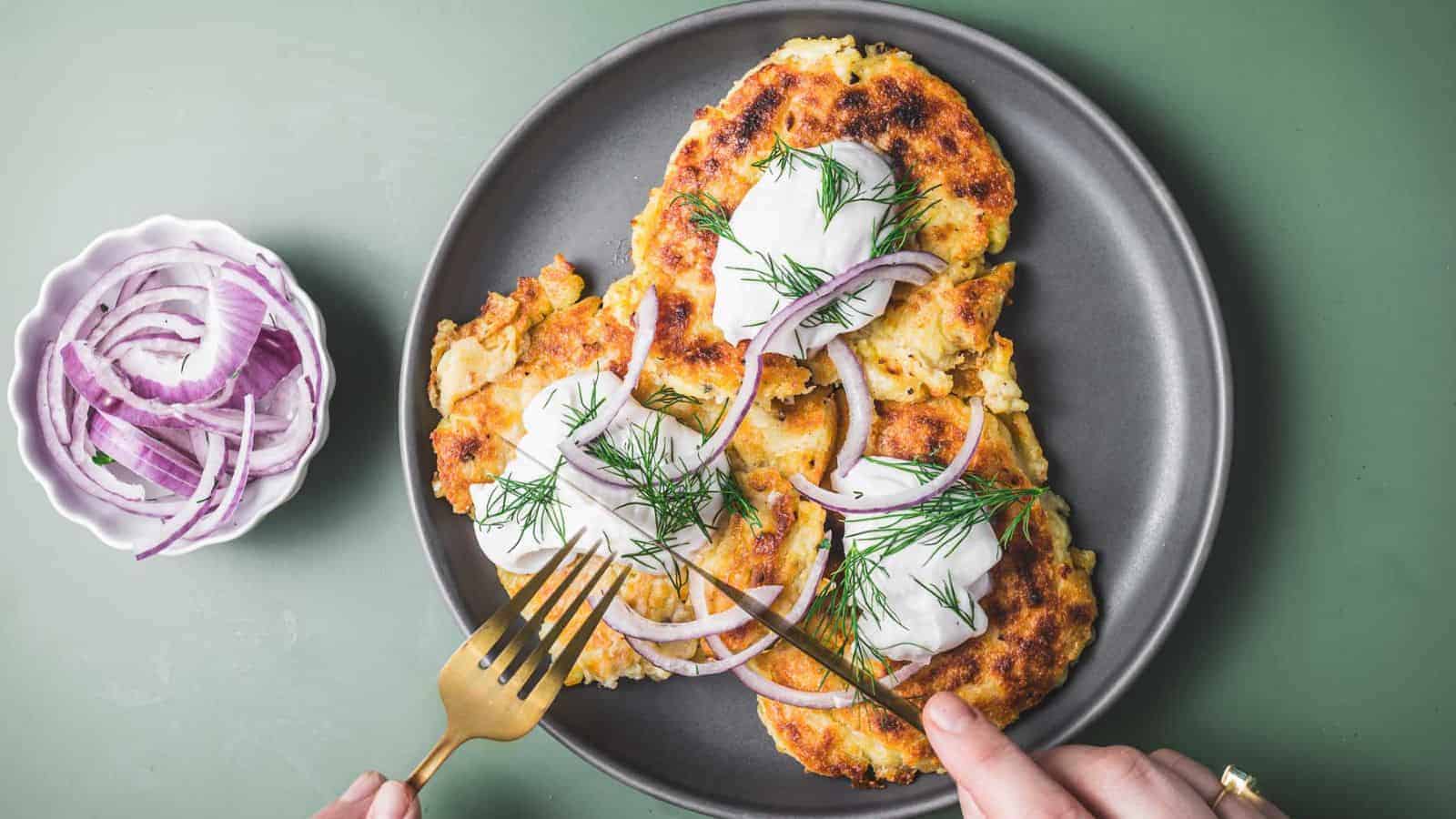 A plate with three golden-brown potato pancakes topped with sour cream, dill, and red onion slices. A hand holds a fork and knife, poised to cut into one pancake. A small bowl of red onion slices is on the side. The background is green.