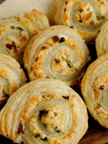 A wooden board holds eight spiral puff pastry pinwheels filled with a cream cheese mixture, topped with small bits of green chili and bacon. A blue and white kitchen towel is partially visible on the left, resting on a marble surface.