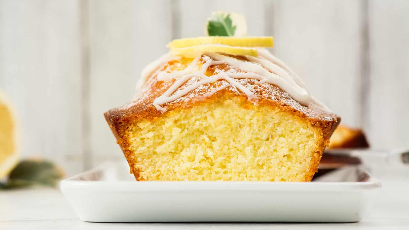 A close-up of a lemon loaf cake with a moist, dense texture, topped with white icing and a lemon slice garnish. The cake is presented on a white rectangular plate against a blurred white background.