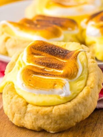 Four lemon meringue cookies with browned meringue tops are on a wooden surface. Three are on a white plate, and one is on a red and white checkered cloth. The cookies have a golden crust, creamy lemon filling, and browned meringue peaks.
