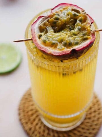 A tall glass filled with yellow juice garnished with half a passion fruit on a skewer sits on a woven coaster. Halves of lime and passion fruit are scattered around on a light surface.