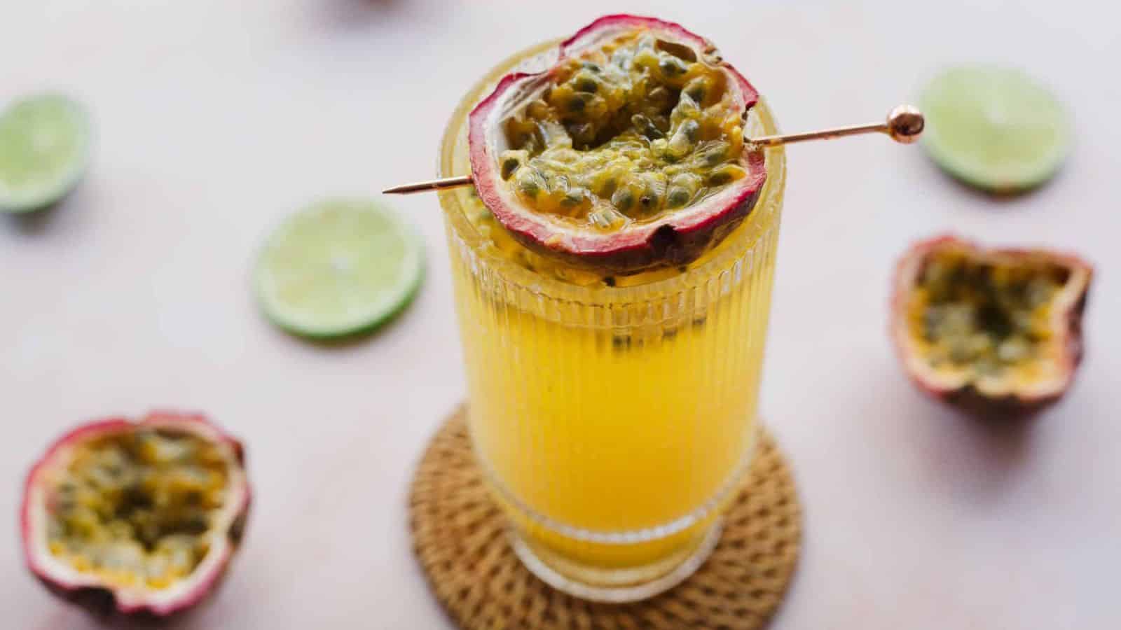 A tall glass filled with yellow juice garnished with half a passion fruit on a skewer sits on a woven coaster. Halves of lime and passion fruit are scattered around on a light surface.