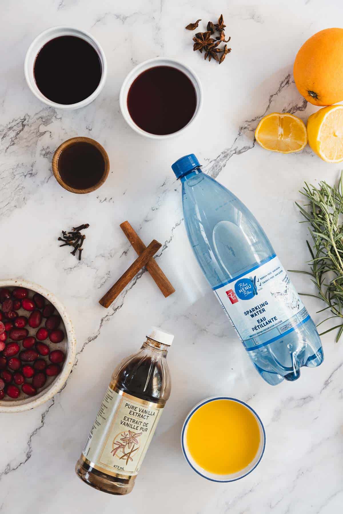 Various ingredients arranged on a marble surface, including two cups with dark liquids, a bottle of sparkling water, a bowl of cranberries, a small cup of brown liquid, lemon halves, an orange slice, vanilla extract, orange juice, cinnamon sticks, and herbs.