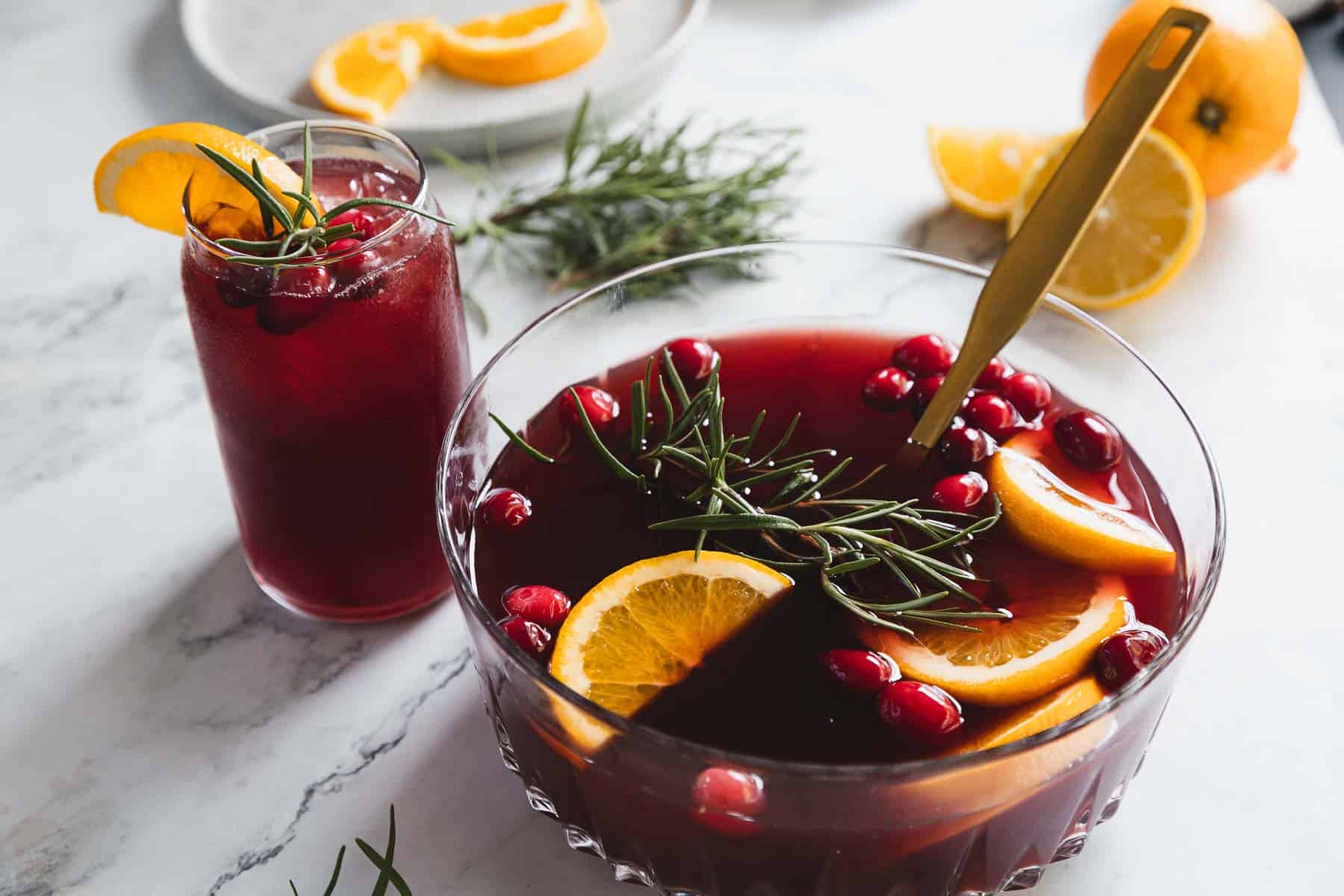 A glass bowl of red punch with cranberries, orange slices, and rosemary, accompanied by a gold ladle. A tall glass filled with the same punch is on the side, garnished with an orange slice and rosemary. Orange slices are in the background.
