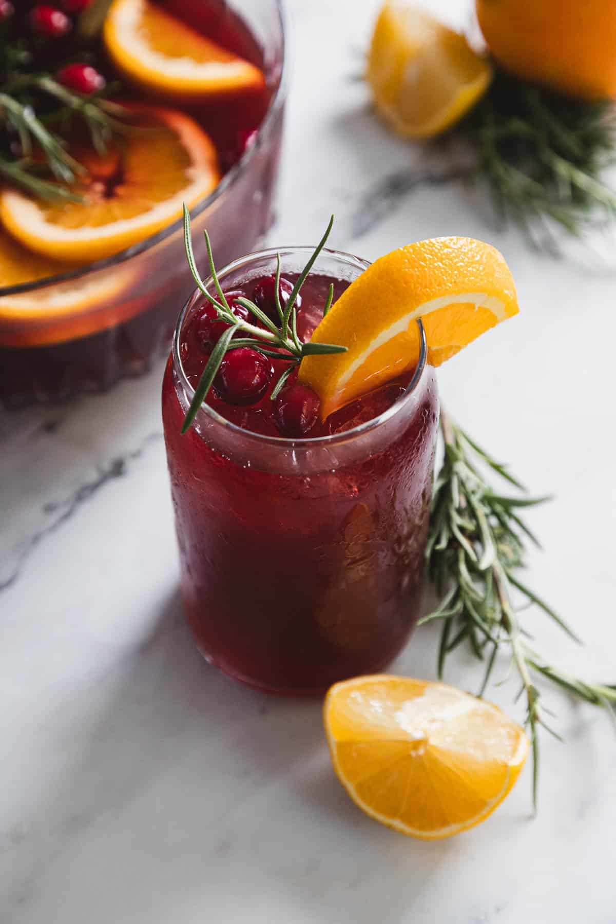 A glass of red beverage garnished with cranberries, a rosemary sprig, and an orange slice sits on a marble surface. A lemon wedge lies nearby, with part of a drink pitcher in the background.