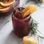 A glass of iced beverage with deep red liquid, garnished with cranberries, a sprig of rosemary, and an orange wedge, is placed on a marble surface. A lemon wedge and rosemary sprigs are nearby, with a partially visible bowl of fruit in the background.