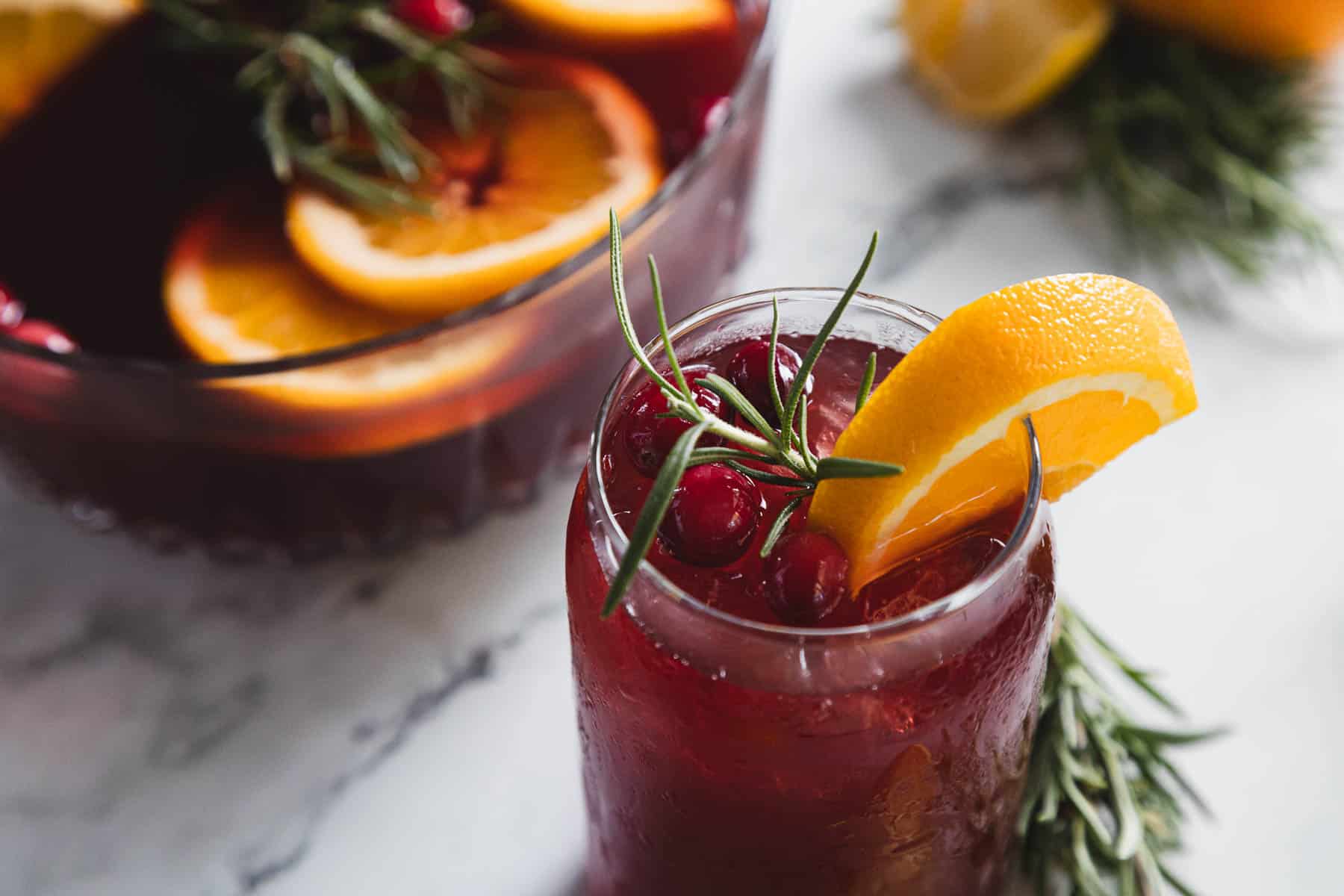 A glass of red sangria garnished with an orange slice, cranberries, and rosemary is in the foreground. In the background, a punch bowl contains more of the same drink with orange slices floating inside. Both are on a marble surface.