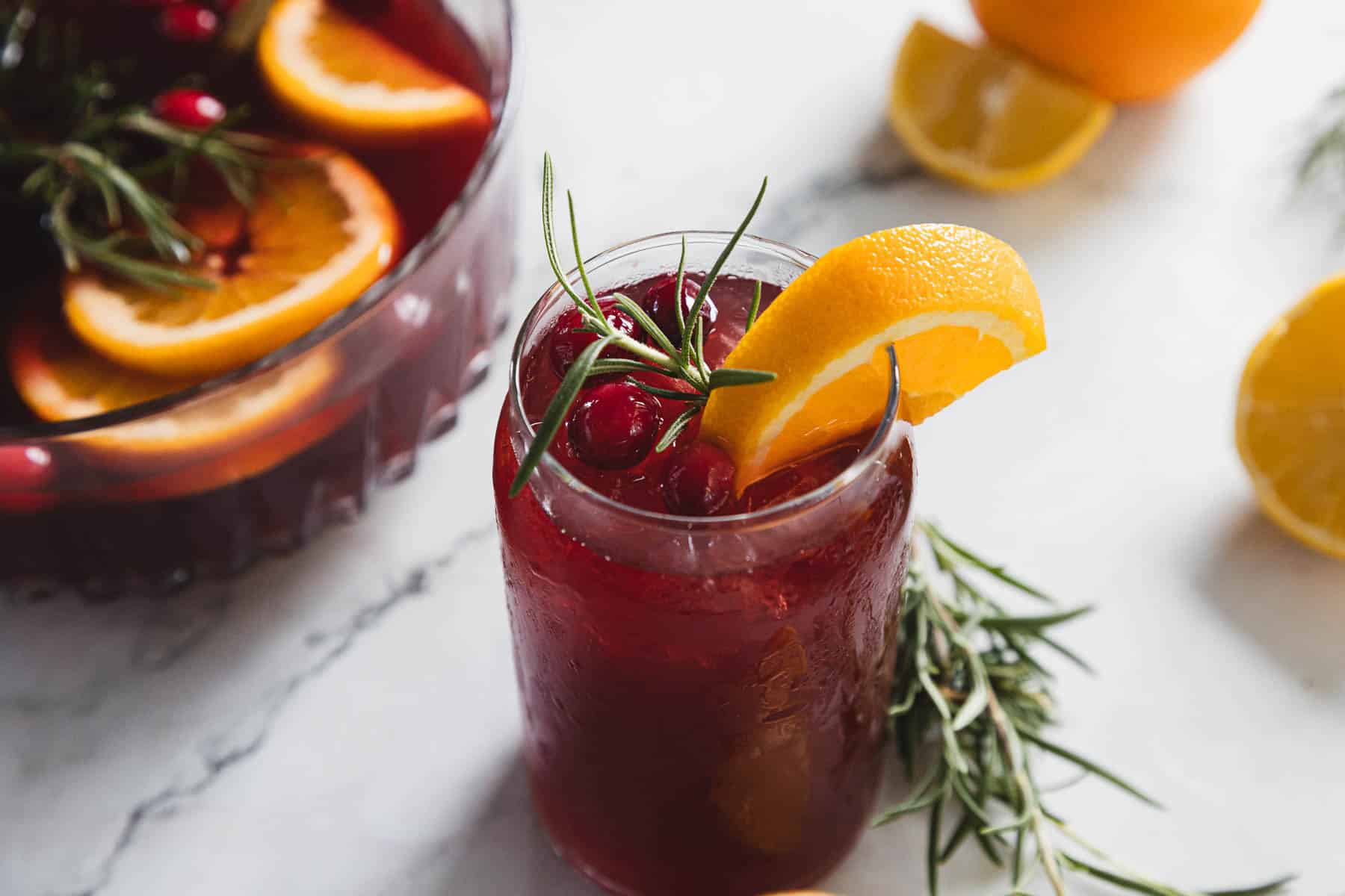 A glass of red punch garnished with an orange slice, cranberries, and a sprig of rosemary sits on a marble surface. A punch bowl with a similar drink and additional orange and lemon slices are in the background.