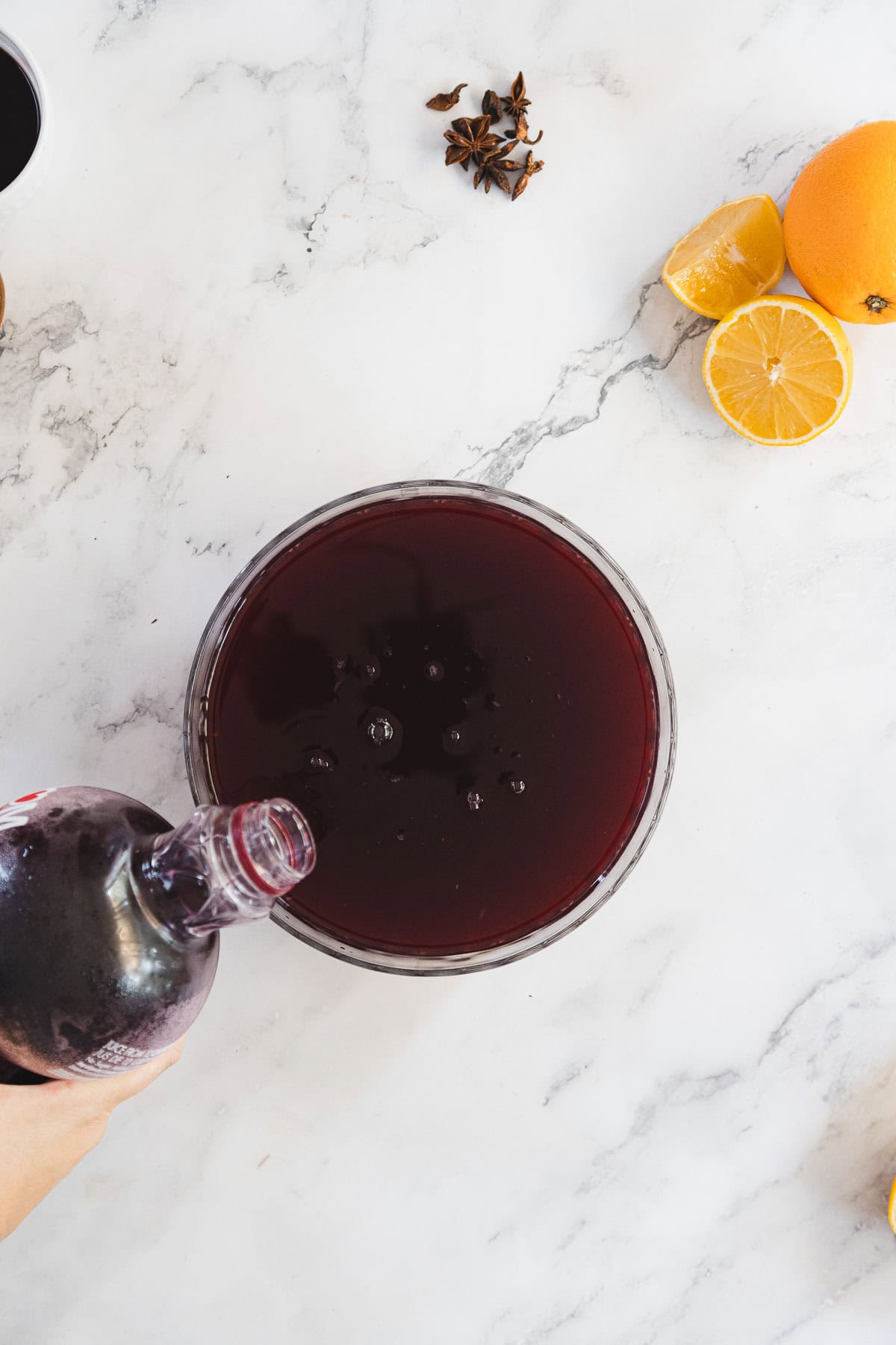 A person pours a dark liquid from a bottle into a large bowl on a marble countertop. Nearby are a sliced lemon, a partially visible orange, and some star anise.