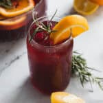 A glass of red punch garnished with orange slices, cranberries, and rosemary sits on a marble surface. A punch bowl is in the background. Orange wedges and rosemary sprigs are scattered around.