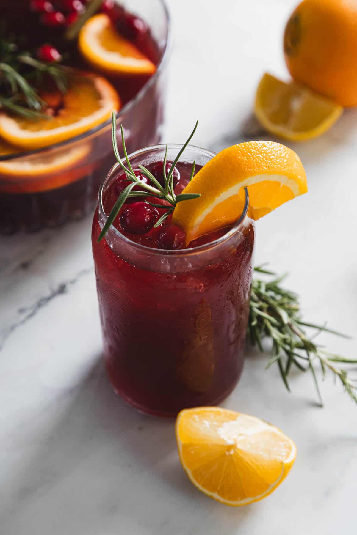 A glass of red punch garnished with orange slices, cranberries, and rosemary sits on a marble surface. A punch bowl is in the background. Orange wedges and rosemary sprigs are scattered around.