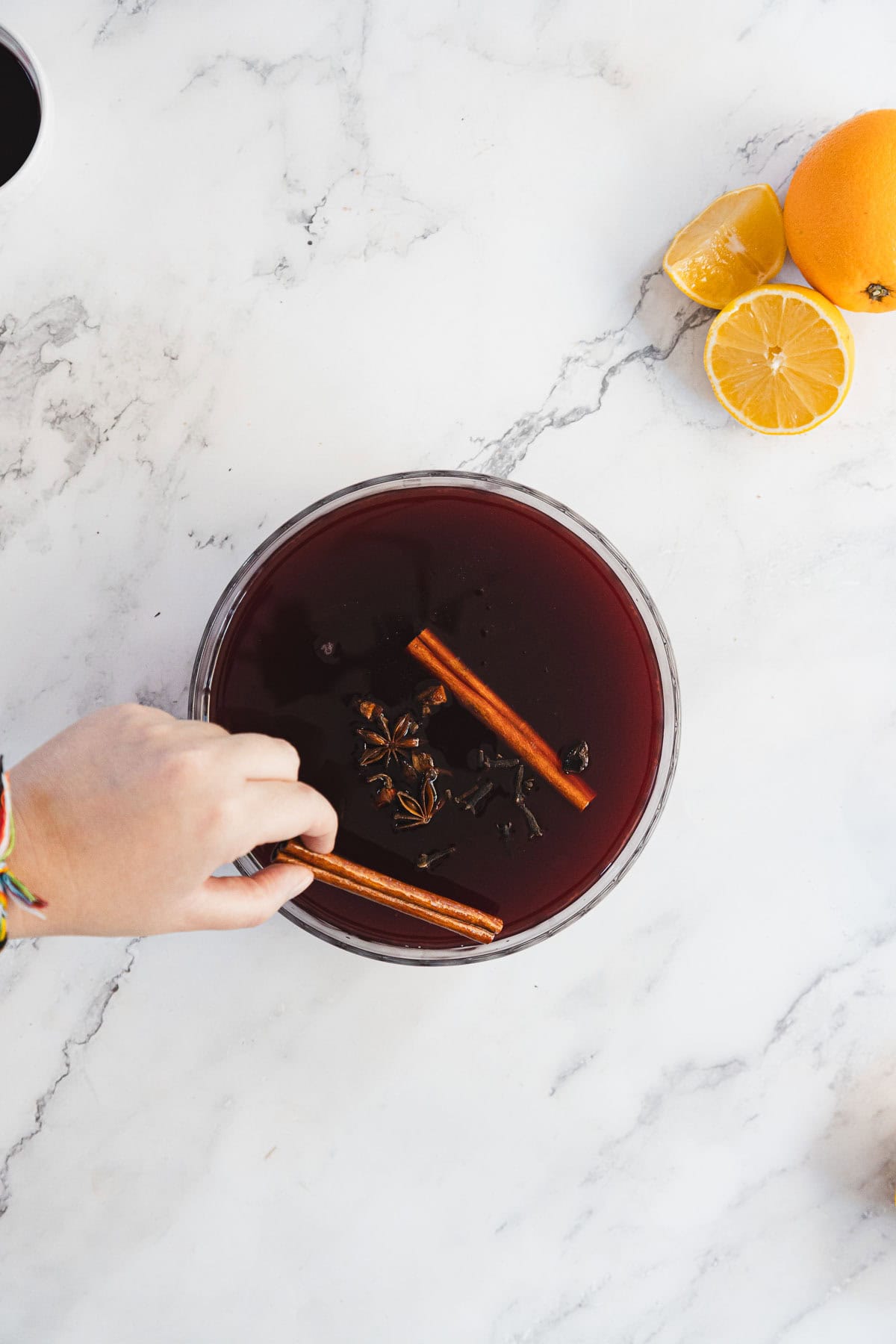 A hand places a cinnamon stick into a bowl of dark red liquid, likely mulled wine, garnished with star anise. On the marble countertop are half an orange and a lemon.