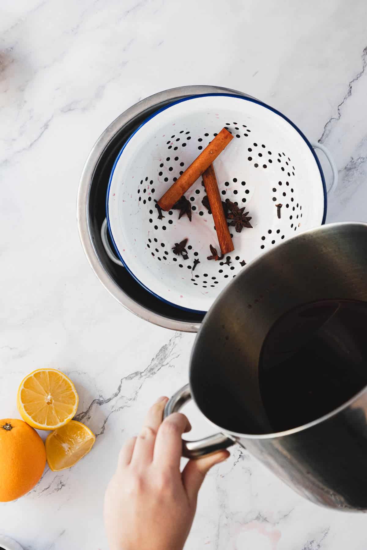 A hand holds a pot over a colander containing two cinnamon sticks and several star anise. Next to the colander are a halved lemon and an orange on a marble countertop.