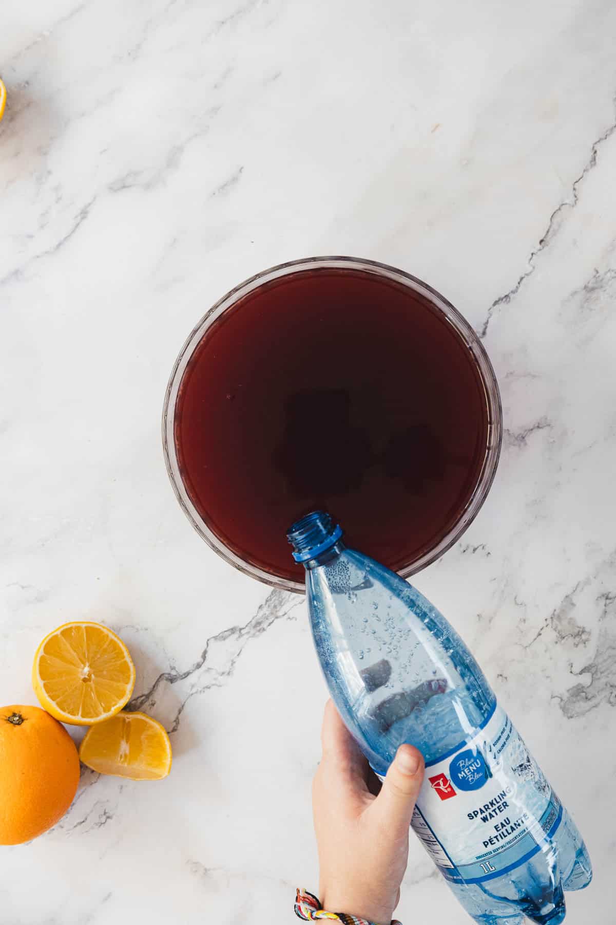 A hand pours sparkling water from a blue plastic bottle into a bowl filled with a dark red liquid. Half an orange and half a lemon are on the marble surface next to the bowl.