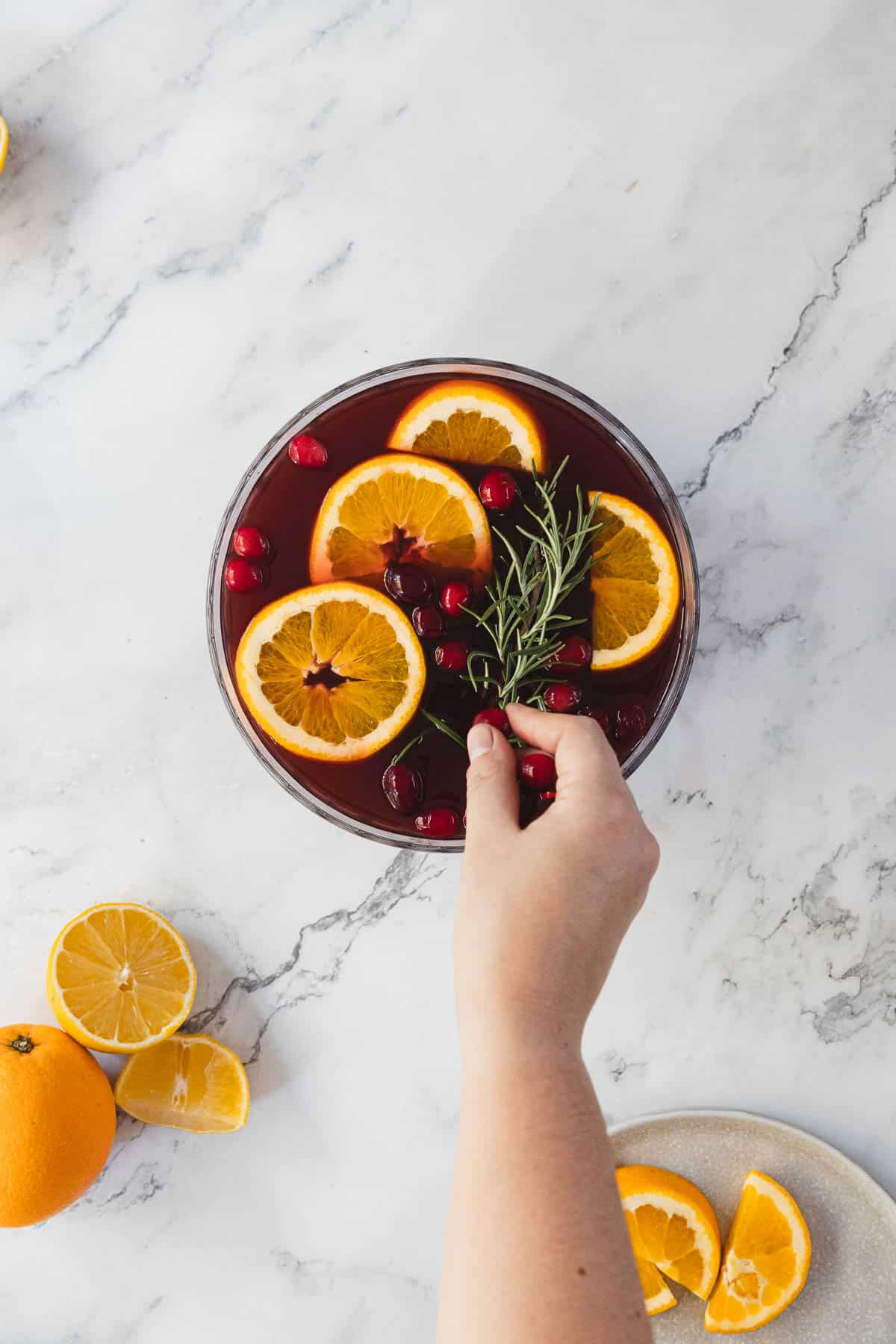 A hand places a sprig of rosemary into a bowl of punch on a marble surface. The punch contains orange slices, cranberries, and rosemary. Nearby are sliced and whole oranges on the table.
