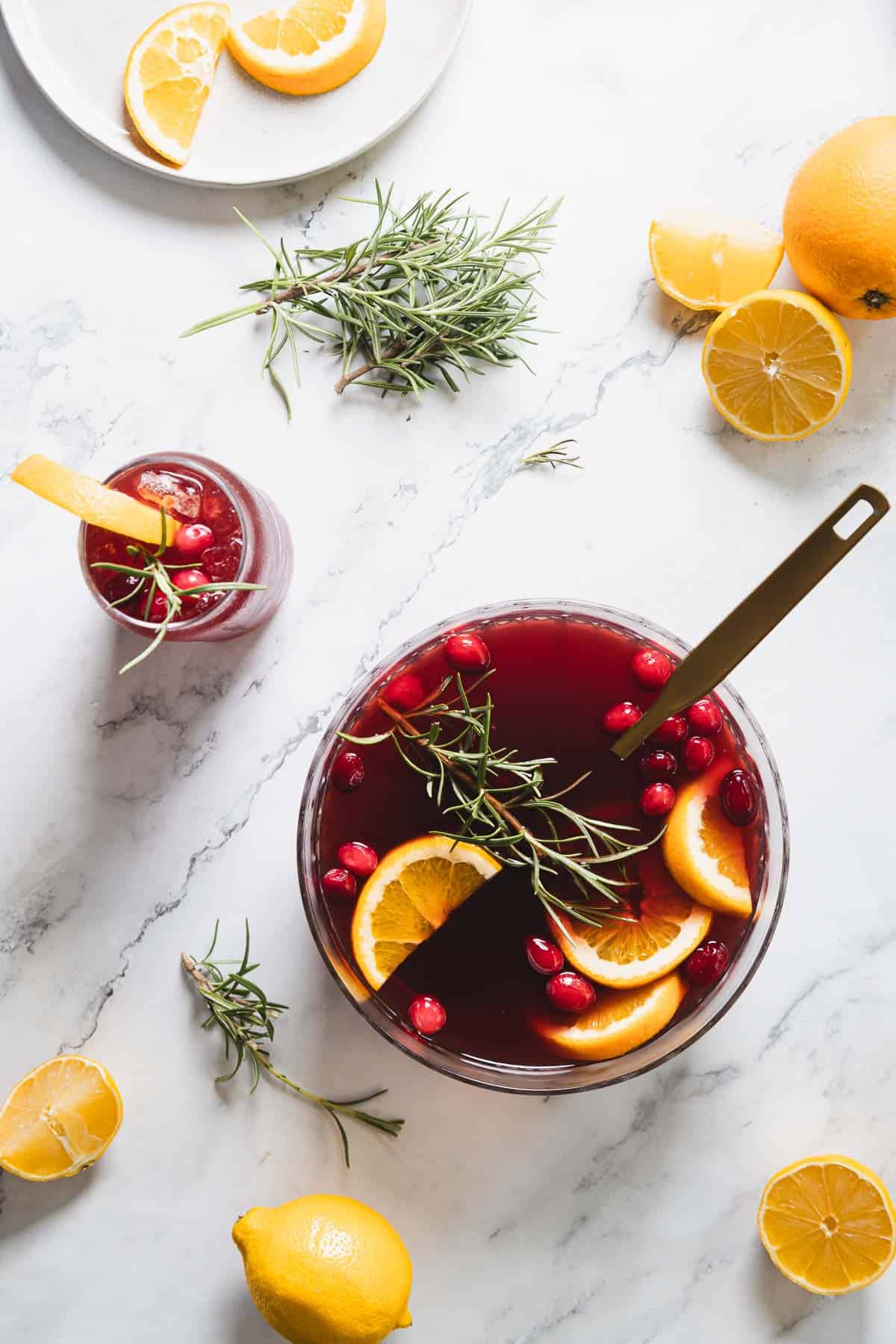 Overhead view of a bowl of red punch with orange slices, cranberries, and rosemary sprigs. A ladle rests inside. A glass of punch with similar garnishes is nearby, surrounded by halved lemons and oranges on a marble surface.